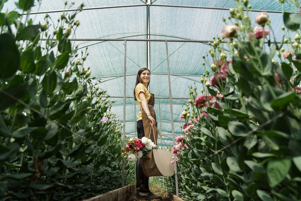 Woman florist walking among flowers in a green house carrying a basket with a fresh bouquet - Fotoğraf, Görsel