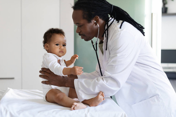 Τσέκαρε το μωρό. Μαύρος παιδίατρος γιατρός που εξετάζει το μικρό βρέφος κατά τη διάρκεια του διορισμού του στην κλινική, Αφροαμερικανός ιατρός με στολή που ελέγχει την υγεία των παιδιών στο νοσοκομείο, κοντινό πλάνο - Φωτογραφία, εικόνα