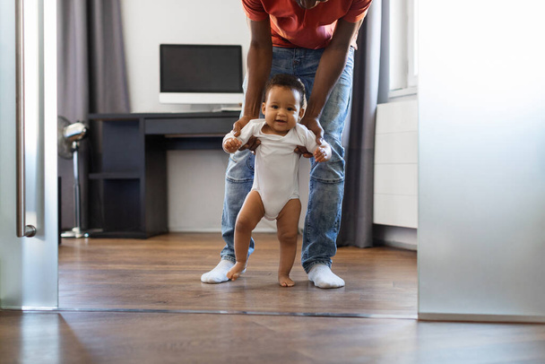 Улюблене афро-американське немовля, що ходить у вітальні з батьками, милий маленький чорношкірий хлопчик або дівчинка робить перші кроки вдома, дбайливий африканський батько-американець, який підтримує свою дитину, затиснутий. - Фото, зображення