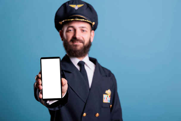 Αεροπλάνο capitan δείχνει smartphone με λευκή λευκή οθόνη γκρο πλαν, τηλέφωνο διαφήμιση προϊόντων mockup με αντίγραφο χώρου. Αεροπλάνο χειριστή κρατώντας τηλέφωνο με κενή οθόνη για διαφημίσεις promo app - Φωτογραφία, εικόνα