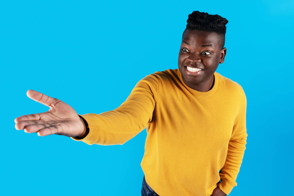 Ενθουσιασμένος Αφροαμερικάνος που απλώνει το χέρι του, κρατώντας αόρατο αντικείμενο στην παλάμη του, κατάπληκτος νεαρός μαύρος που δείχνει κάτι στο χέρι του ενώ στέκεται απομονωμένος σε μπλε φόντο, αντιγράφει χώρο - Φωτογραφία, εικόνα