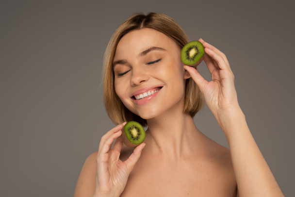 gelukkig jong vrouw met gesloten ogen houden kiwi helften geïsoleerd op grijs  - Foto, afbeelding