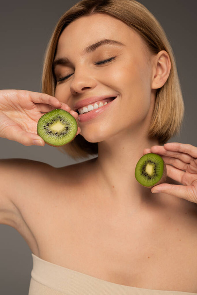 cheerful young woman with closed eyes holding kiwi fruit halves isolated on grey - Zdjęcie, obraz
