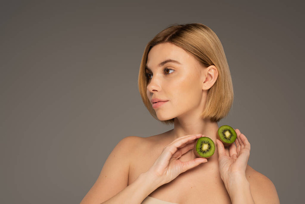 jonge vrouw met blote schouders houden kiwi fruit en weg te kijken geïsoleerd op grijs - Foto, afbeelding