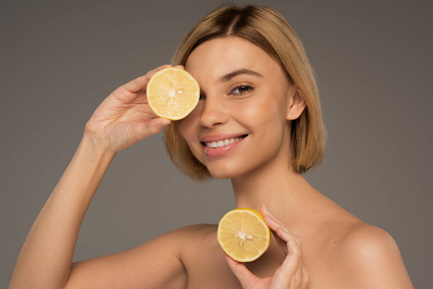 positive woman with bare shoulders holding lemon halves isolated on grey  - Foto, Bild