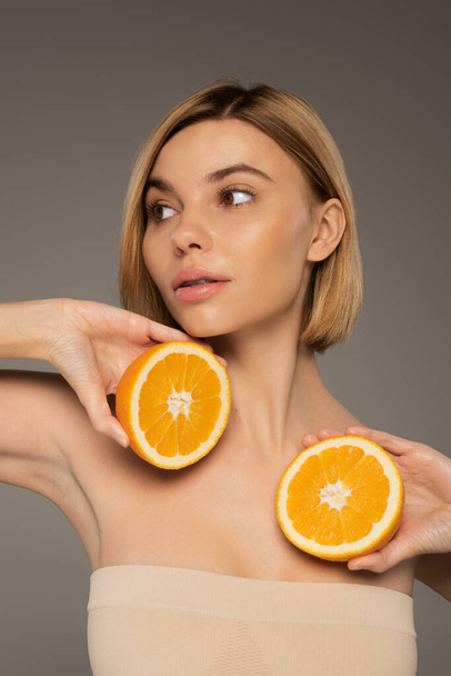 pretty young woman with bare shoulders holding orange halves isolated on grey  - Zdjęcie, obraz