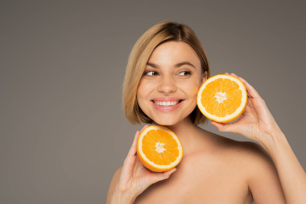 cheerful woman with bare shoulders holding orange halves isolated on grey  - Foto, immagini