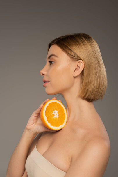πλαϊνή άποψη της όμορφης νεαρής γυναίκας με γυμνούς ώμους κρατώντας πορτοκαλί απομονωμένο σε γκρι  - Φωτογραφία, εικόνα