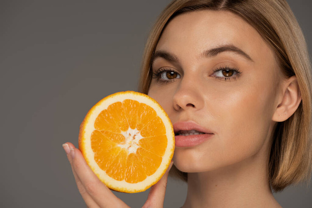 portrait of young blonde woman holding juicy orange half isolated on grey  - Foto, afbeelding