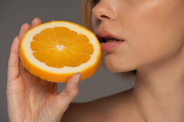 cropped view of young woman holding juicy orange half near soft lips isolated on grey  - Fotó, kép