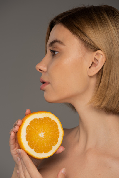 profile of young blonde woman holding juicy orange half isolated on grey  - Foto, immagini