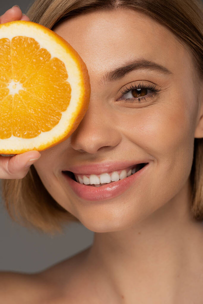 portrait of cheerful blonde woman holding juicy orange half near face isolated on grey  - 写真・画像