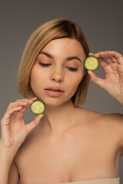 blonde woman with naked shoulders holding sliced fresh cucumbers isolated on grey  - Foto, Imagen