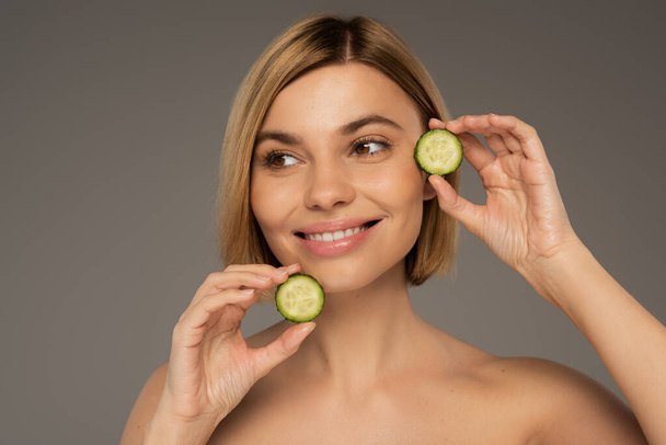 cheerful young woman with naked shoulders holding sliced fresh cucumbers isolated on grey  - Foto, immagini