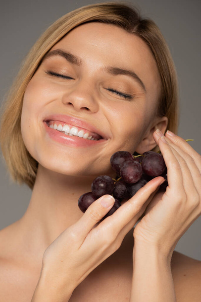 vrolijke jonge vrouw met verse druiven en glimlachen geïsoleerd op grijs  - Foto, afbeelding