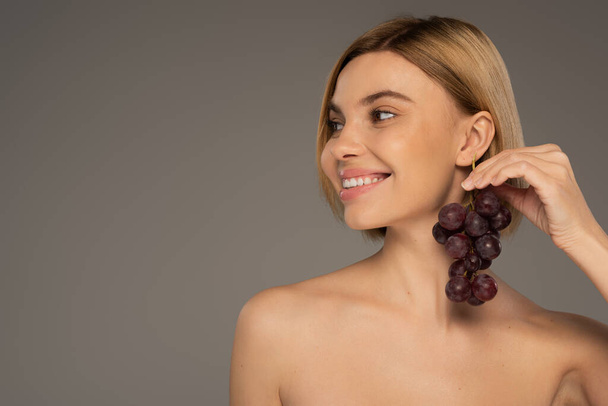 happy young woman with bare shoulders holding ripe grapes near ear isolated on grey  - Foto, Imagen