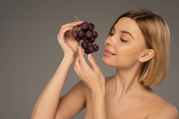 vrolijke jonge vrouw met blote schouders houden rijpe druiven geïsoleerd op grijs  - Foto, afbeelding