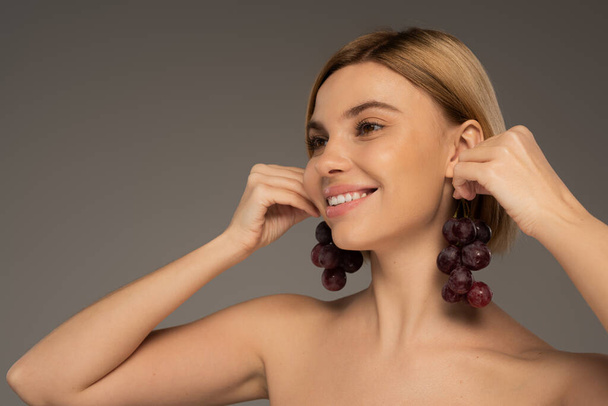 vrolijke jonge vrouw met blote schouders houden smakelijke druiven in de buurt van oren geïsoleerd op grijs  - Foto, afbeelding