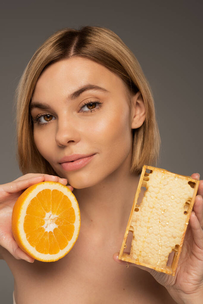 blond jong vrouw holding honingraat en oranje terwijl kijken naar camera geïsoleerd op grijs  - Foto, afbeelding