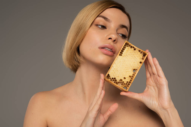 blond jong vrouw holding houten frame met zoete honing terwijl weg te kijken geïsoleerd op grijs  - Foto, afbeelding