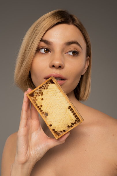 blonde young woman holding wooden frame with sweet honey near face isolated on grey  - Fotó, kép