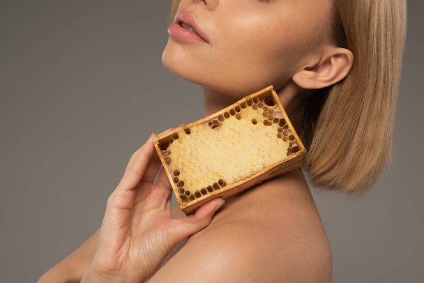 bijgesneden uitzicht van blonde jonge vrouw met houten frame met zoete honing geïsoleerd op grijs  - Foto, afbeelding