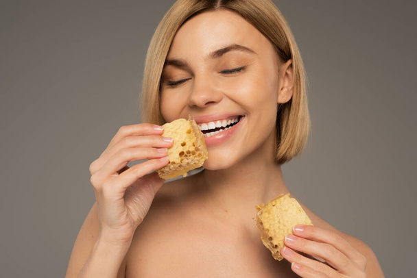 joyful woman with bare shoulders holding pieces of sweet honeycomb isolated on grey - Фото, изображение