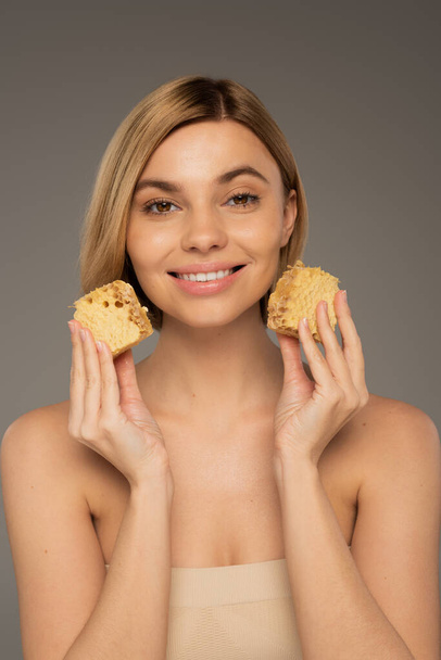 pleased young woman holding sweet honeycombs isolated on grey - Foto, immagini