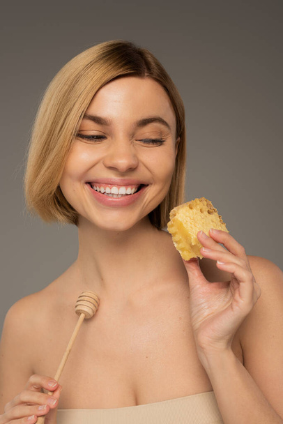 pleased young woman with naked shoulders holding sweet honeycomb and wooden dipper isolated on grey - Foto, Imagen