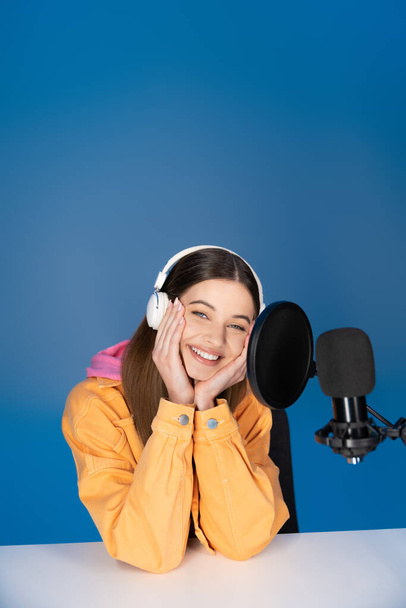 Smiling teenager in headphones sitting near studio microphone isolated on blue  - 写真・画像