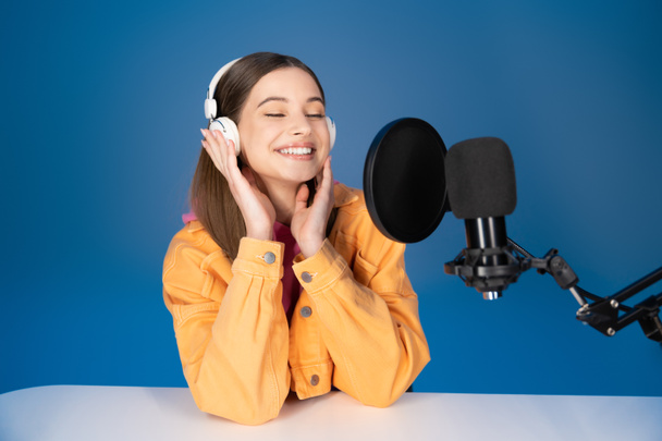 Ευτυχισμένος έφηβος με ακουστικά κάθεται κοντά στο μικρόφωνο στούντιο απομονώνονται σε μπλε  - Φωτογραφία, εικόνα