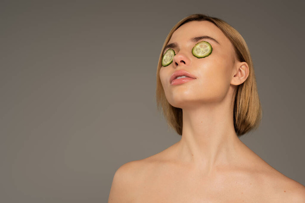 blonde young woman with naked shoulders and fresh cucumbers on eyes isolated on grey  - Foto, imagen