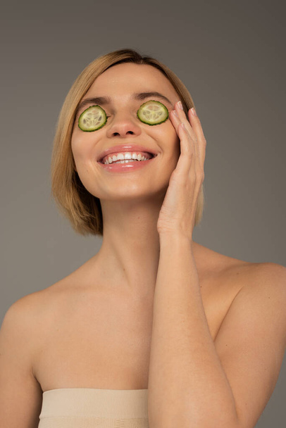 joyful young woman with naked shoulders and fresh cucumbers on eyes isolated on grey  - Foto, Imagen