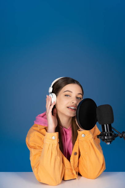 Брюнетка подросток касаясь наушников и говорить возле студии микрофон изолирован на синий  - Фото, изображение