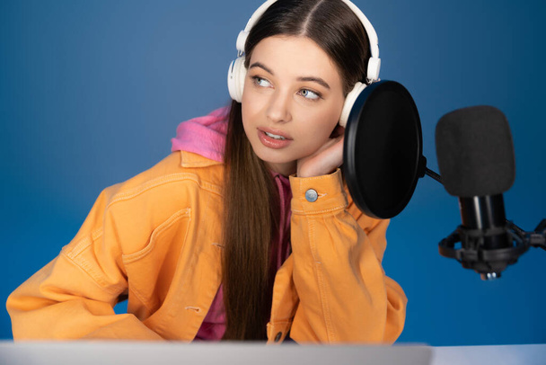 Teenager in headphones looking away near studio microphone isolated on blue  - Photo, image