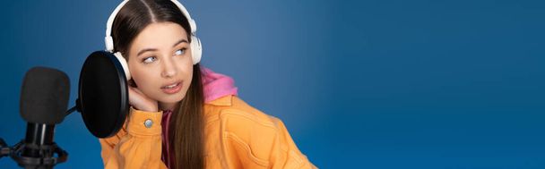 Teen girl in wireless headphones looking away near studio microphone isolated on blue, banner  - Foto, Imagem