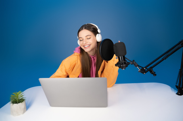 Улыбающаяся девушка в наушниках с помощью ноутбука рядом с микрофоном на столе изолированы на синий  - Фото, изображение