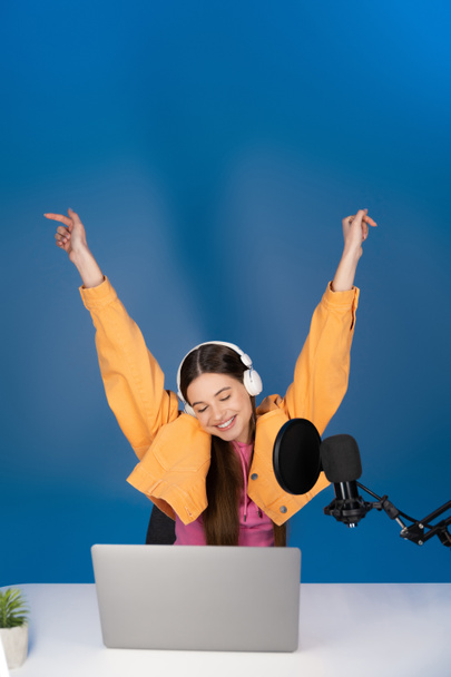 Удовлетворенный подросток в наушниках сидит с поднятыми руками возле микрофона и ноутбука на синем фоне  - Фото, изображение