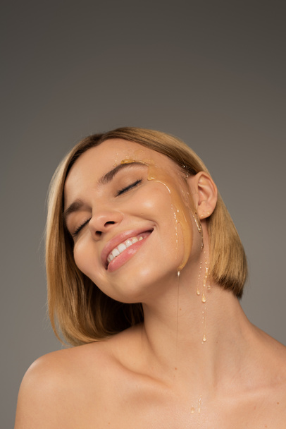 gelukkig jong vrouw met zoet honing op wang glimlachen geïsoleerd op grijs  - Foto, afbeelding