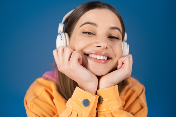 Portrait of cheerful teenager in headphones looking at camera isolated on blue  - Valokuva, kuva