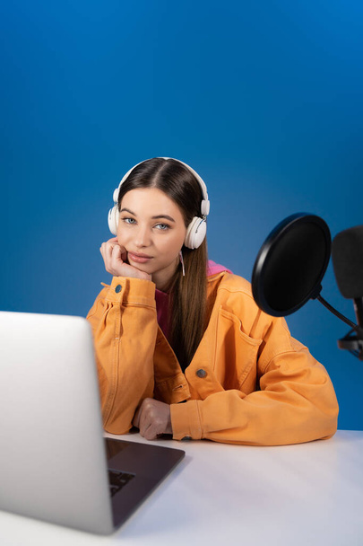 Девушка-брюнетка в наушниках смотрит в камеру рядом с размытым ноутбуком и студийным микрофоном, изолированным на синем  - Фото, изображение