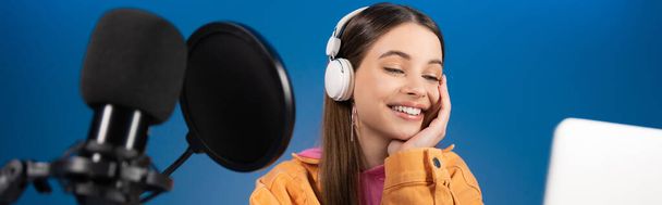 Χαρούμενος έφηβος με ακουστικά να κάνει podcast κοντά σε φορητό υπολογιστή και μικρόφωνο στούντιο απομονωμένο σε μπλε, banner  - Φωτογραφία, εικόνα