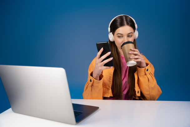 Teenager in headphones using smartphone and drinking takeaway coffee near laptop isolated on blue  - Fotó, kép