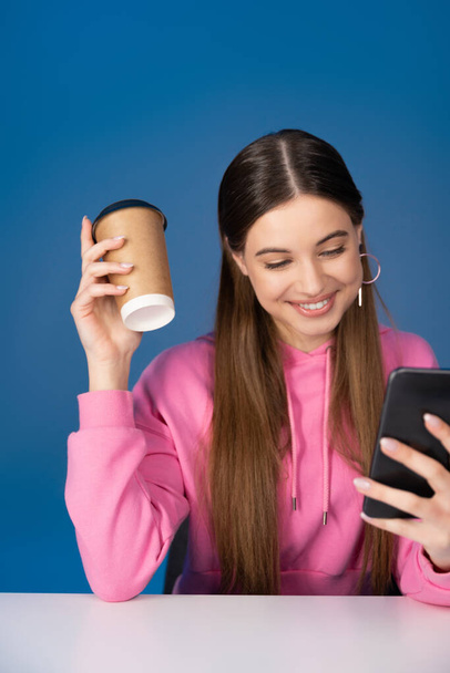 Positiivinen teini tyttö tilalla kahvia mennä ja käyttää älypuhelin eristetty sininen  - Valokuva, kuva
