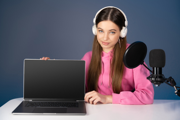 Teen podcaster in headphones sitting near laptop and microphone isolated on blue  - Photo, image