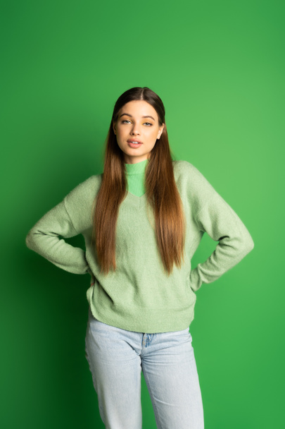 Красивая девушка-подросток в свитере держит за руки бедра на зеленом фоне - Фото, изображение