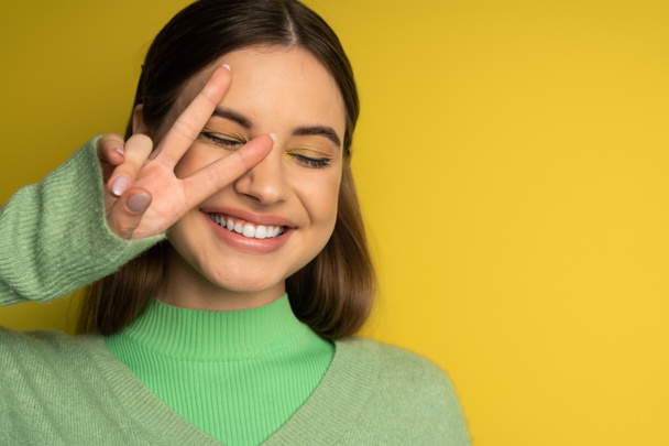 Позитивная девочка-подросток в свитере показывает знак мира на желтом фоне  - Фото, изображение