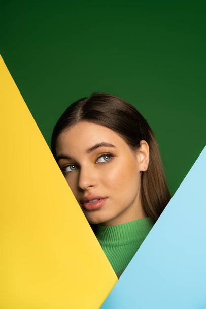 Brunette teenager in turtleneck posing near colorful paper isolated on green  - Foto, imagen