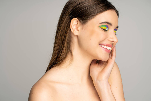 Smiling teen model with colorful visage and naked shoulders touching face isolated on grey  - Valokuva, kuva