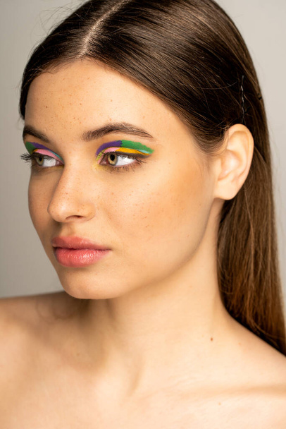 Brunette teen model with freckles and colorful eyeshadows looking away isolated on grey  - Zdjęcie, obraz